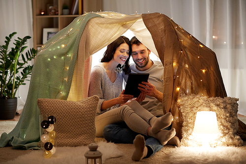 leisure, hygge, technology and people concept - happy couple with tablet pc computer in kids tent at home