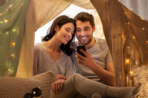leisure, hygge, technology and people concept - happy couple with smartphone in kids tent at home