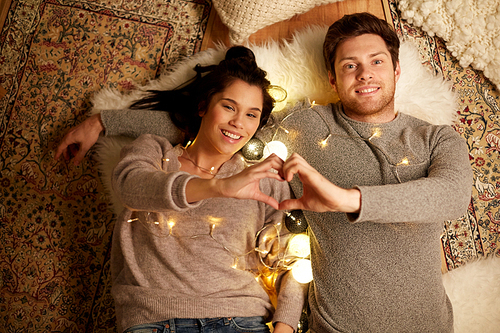 leisure, hygge and people concept - happy couple with garland lying on floor at home and making hand heart gesture
