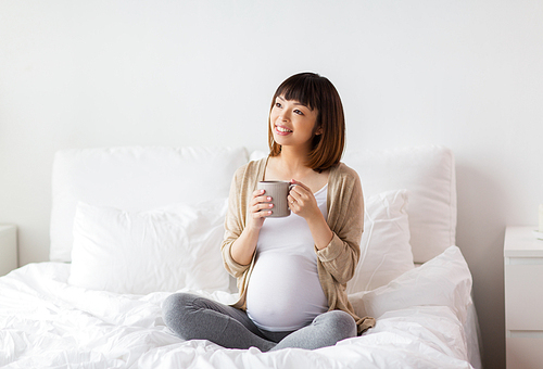 pregnancy, rest, people and expectation concept - happy pregnant asian woman with cup drinking tea in bed at home bedroom