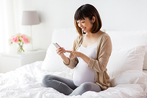pregnancy, technology and people concept - happy pregnant asian woman with smartphone in bed at home