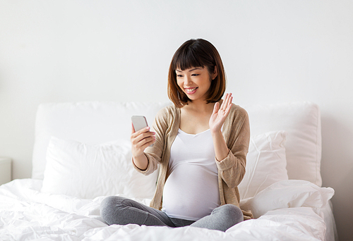 pregnancy, technology and people concept - happy pregnant asian woman taking selfie by smartphone in bed at home and waving hand