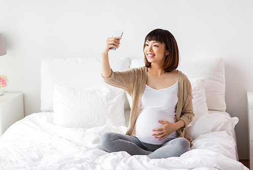 pregnancy, technology and people concept - happy pregnant asian woman taking selfie by smartphone in bed at home