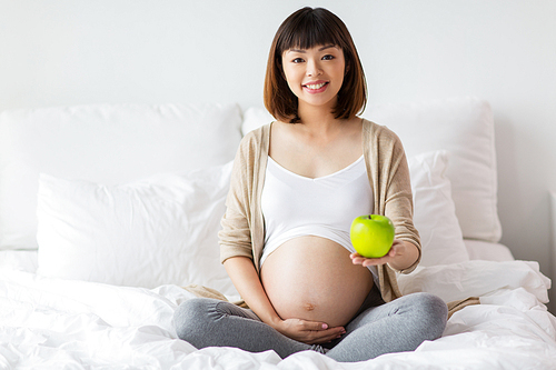 pregnancy, people and healthy eating concept - happy pregnant asian woman sitting in bed at home with green apple