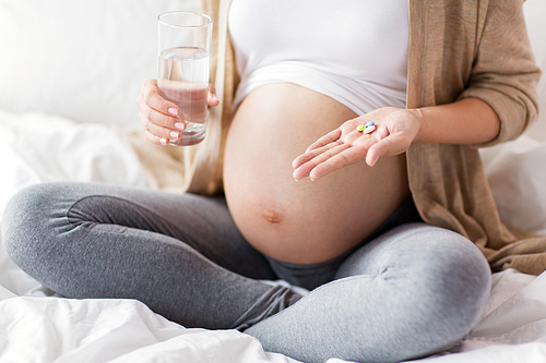 pregnancy, people and health care concept - close up of pregnant woman sitting in bed at home with pills and glass of water