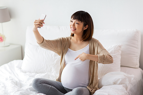 pregnancy, technology and people concept - happy pregnant asian woman taking selfie by smartphone in bed at home and pointing finger to her belly