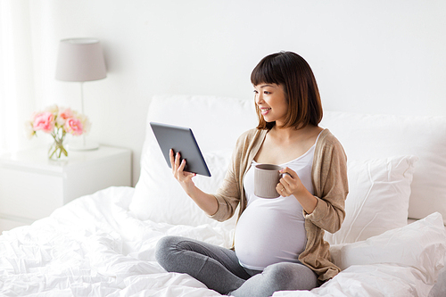 pregnancy, technology, people and expectation concept - happy pregnant asian woman with tablet pc computer drinking tea in bed at home