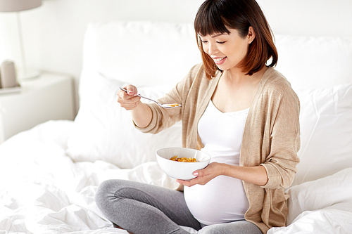 pregnancy, people and breakfast concept - happy pregnant asian woman eating cereal flakes in bed at home bedroom