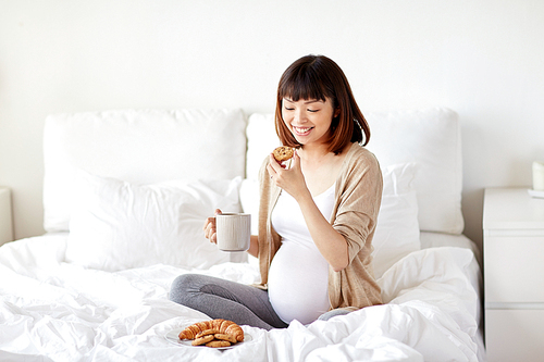 pregnancy, rest, people and breakfast concept - happy pregnant asian woman with cup drinking tea and eating cookie in bed at home bedroom