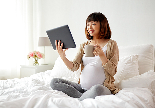 pregnancy, technology, people and expectation concept - happy pregnant asian woman with tablet pc computer drinking tea in bed at home