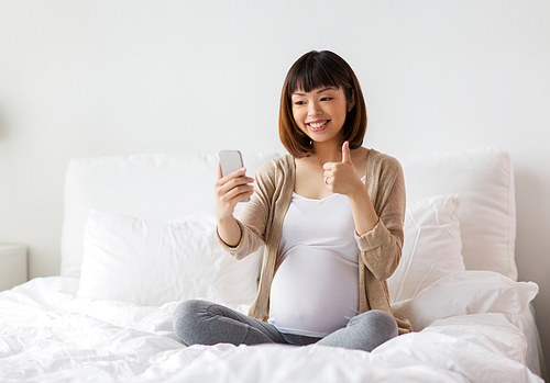 pregnancy, technology and people concept - happy pregnant asian woman taking selfie by smartphone in bed at home and showing thumbs up