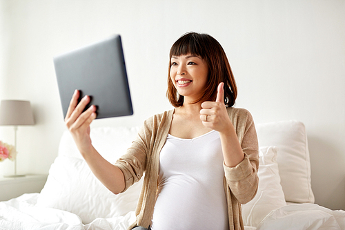pregnancy, technology and people concept - happy pregnant asian woman with tablet pc computer having video call and showing thumbs up in bed at home