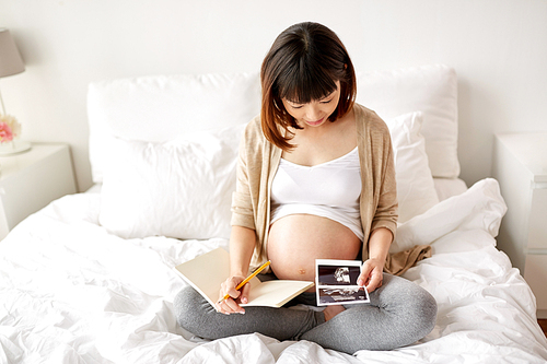 pregnancy and people concept - happy pregnant asian woman with fetal ultrasound image and notebook or diary in bed at home
