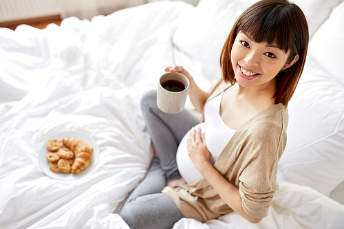 pregnancy, rest, people and breakfast concept - happy pregnant asian woman with cup drinking tea in bed at home bedroom