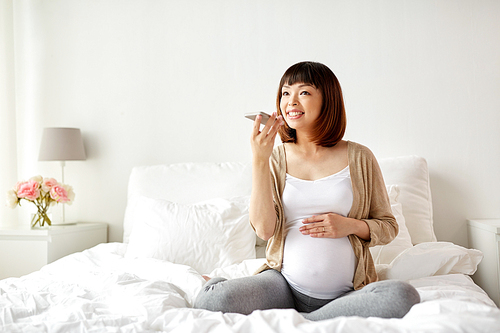 pregnancy, motherhood, technology, people and expectation concept - happy pregnant asian woman using voice command recorder or calling on smartphone at home