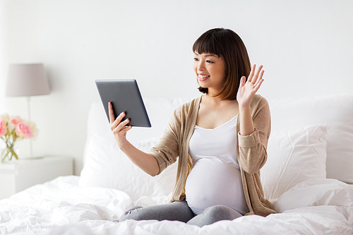 pregnancy, technology and people concept - happy pregnant asian woman with tablet pc computer having video call and waving hand in bed at home