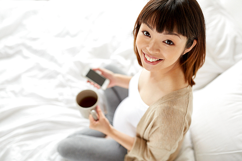 pregnancy, rest, people and expectation concept - happy pregnant asian woman with cup of tea and smartphone in bed at home bedroom