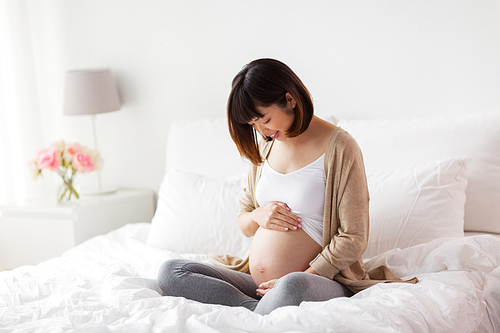 pregnancy, people and maternity concept - happy pregnant asian woman sitting in bed at home bedroom and looking at her bare belly