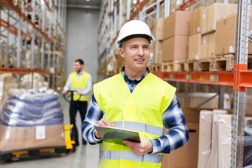 logistic business, shipment and people concept - smiling male worker or supervisor with clipboard in reflective safety vest at warehouse