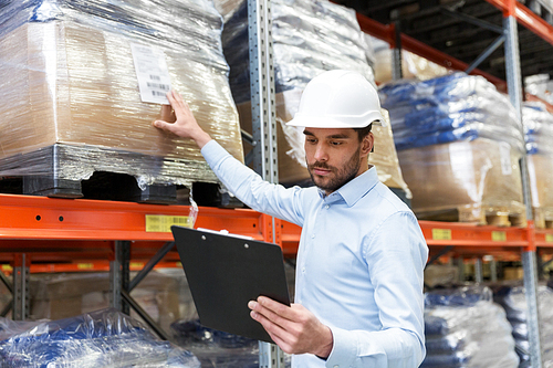 logistic business, shipment and people concept - businessman in helmet with clipboard checking goods at warehouse