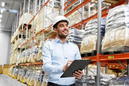 logistic business, shipment and people concept - businessman in helmet with clipboard checking goods at warehouse