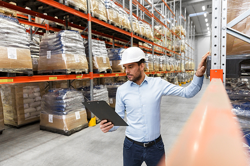 logistic business, shipment and people concept - businessman in helmet with clipboard checking goods at warehouse