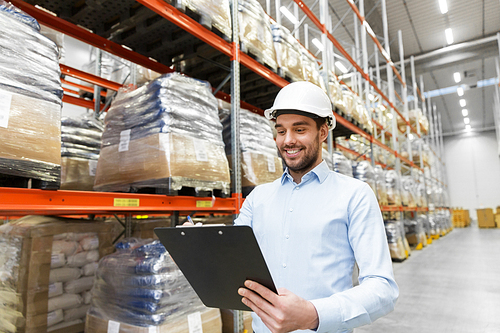 logistic business, shipment and people concept - businessman in helmet with clipboard checking goods at warehouse