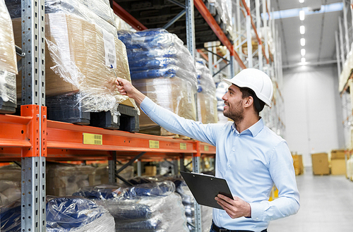 wholesale, logistic, business, export and people concept - man or manager in helmet with clipboard checking goods at warehouse