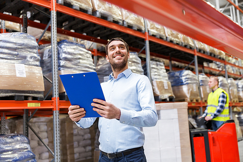 logistic business, shipment and people concept concept - happy businessman with clipboard at warehouse