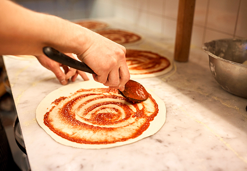 food, culinary, italian cuisine, people and cooking concept - cook with spoon applying tomato sauce to raw pizza dough at pizzeria