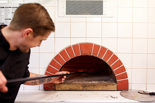 food cooking, culinary and people concept - cook or baker with peel placing pizza to oven at pizzeria