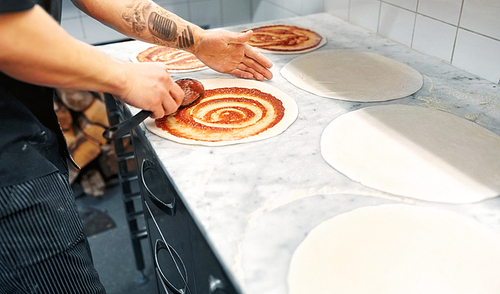 food cooking, culinary and people concept - cook or baker with spoon applying tomato sauce to raw pizza dough at pizzeria