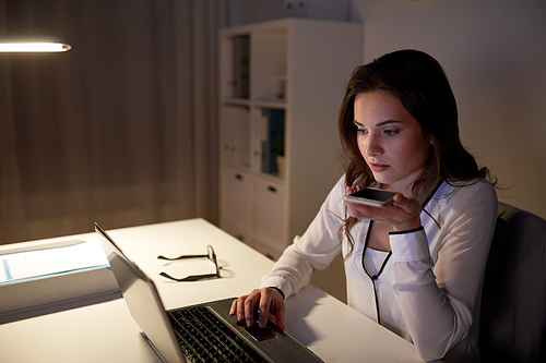 business, technology, overwork, deadline and people concept - woman with laptop and smartphone using voice command recorder at night office