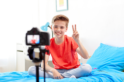 blogging, technology and people concept - happy smiling boy or . with camera recording video at home and showing peace hand sign