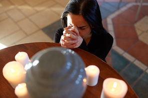 people and mourning concept - sad woman with cinerary urn and candles praying at funeral in church