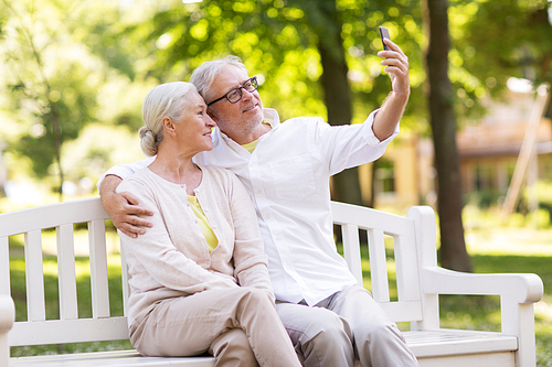 technology, relationship and old people concept - happy senior couple with smartphone taking selfie and hugging at summer park