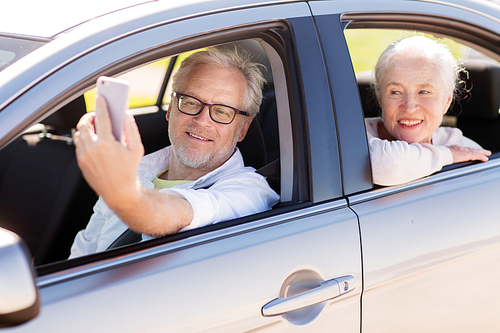 road trip, travel and old people concept - happy senior couple driving in car and taking selfie by smartphone