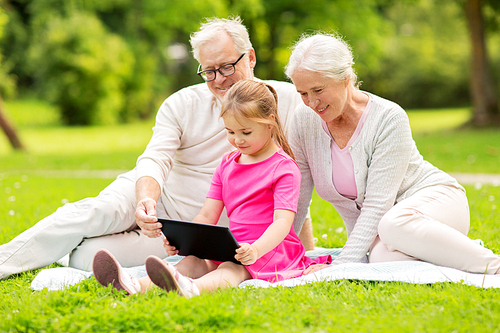 family, generation and technology concept - happy smiling grandmother, grandfather and little granddaughter with tablet pc computer sitting on blanket at park