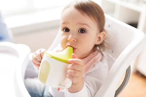 childhood and people concept - little baby drinking from spout cup sitting in highchair at home