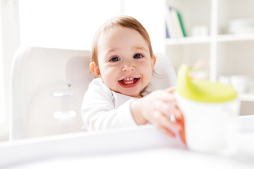 childhood and people concept - happy smiling little baby drinking from spout cup sitting in highchair at home