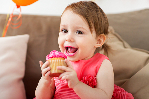 childhood, people and celebration concept - happy baby girl eating cupcake on birthday party at home