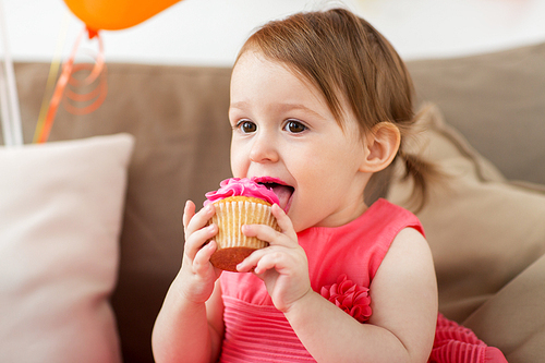 childhood, people and celebration concept - happy baby girl eating cupcake on birthday party at home