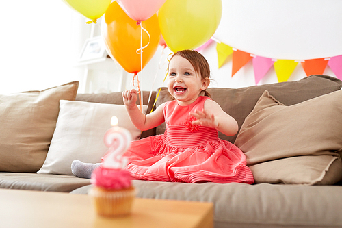 childhood, holidays and people concept - happy baby girl on birthday party at home with air balloons, garland and cupcake with candle