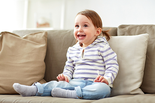 childhood, emotions and people concept - happy smiling baby girl sitting on sofa at home