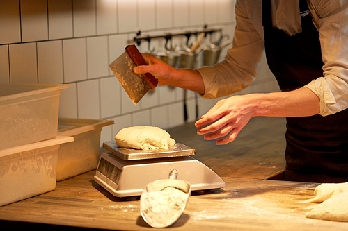 food cooking, baking and people concept - chef or baker with bench cutter portioning and weighing dough on scale at bakery