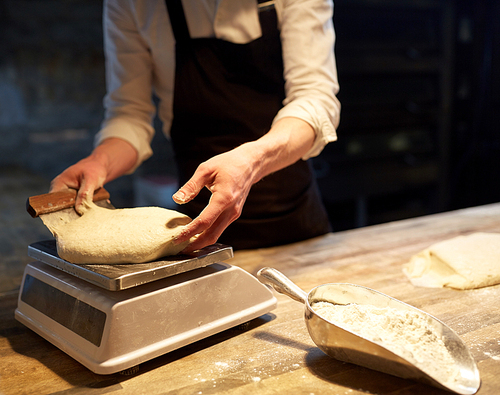 food cooking, baking and people concept - chef or baker with bench cutter portioning and weighing dough on scale at bakery