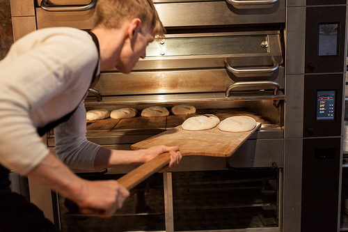 food, cooking and baking concept - baker with peel putting yeast dough into bread oven at bakery kitchen