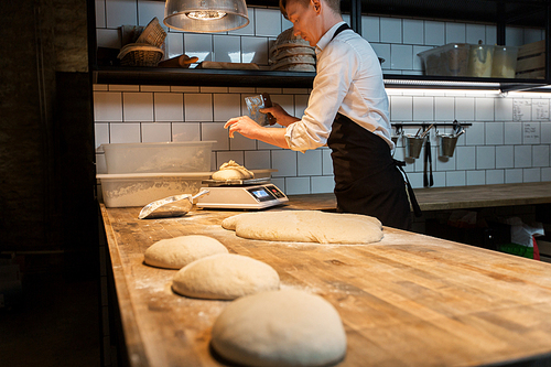 food cooking, baking and people concept - chef or baker with bench cutter portioning and weighing dough on scale at bakery kitchen