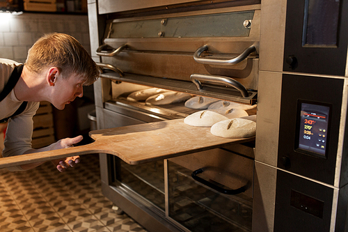 food, cooking and baking concept - baker with peel putting yeast dough into bread oven at bakery kitchen