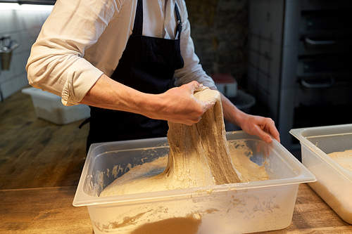 food cooking, baking and people concept - baker kneading dough and making bread at bakery kitchen